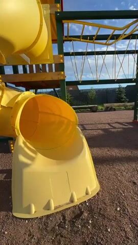 A young child wearing an orange jacket and light-colored pants is playing on a yellow slide at a playground. The playground structure is yellow and green with a blue sky and white clouds in the background. The ground of the playground is covered in reddish-brown gravel. The child slides down the tube slide and then walks away from it. The camera pans slightly to the right as the child moves.