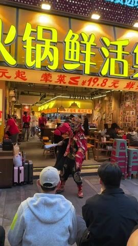 The video shows a street performance in front of a restaurant. The performer is wearing a red and black traditional Chinese costume, a mask, and tall boots. He is holding a blue fan and a small sword, and he is gesturing with both. Several people are watching the performance, including two men in the foreground who are seated. Behind the performer is the entrance to the restaurant, which is brightly lit and has yellow signage in Chinese characters. There are people inside the restaurant, some of whom appear to be employees wearing red shirts. Luggage sits to the left of the people watching the performance.