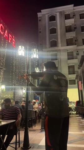 The video shows a man playing a violin in an outdoor restaurant setting. The man is standing near a lamp post, facing away from the camera, and is wearing a dark shirt and dark pants with an orange stripe. He is playing the violin with his right hand using the bow. Behind him, people are seated at tables, dining at the outdoor cafe, and there is a building with the words "LM CAFE" in large red neon letters. The cafe is adorned with strings of small lights, creating a festive atmosphere. Across from the cafe is a taller, modern-looking building with visible balconies and windows. The sky is dark, suggesting it is nighttime.