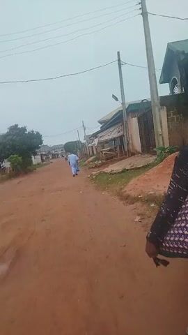 A woman with dark hair, wearing a dark patterned top and dark pants, walks away from the camera down a dirt road. She carries a patterned tote bag over her right shoulder and a dark plastic bag in her left hand. The road is lined with buildings and some vegetation. Another person, dressed in a light blue garment, walks further down the road in the same direction. The sky is overcast.