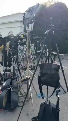 The video shows a collection of professional video cameras and lighting equipment set up outside a white building, likely for news coverage. The cameras are mounted on tripods, and there are lighting fixtures on stands. Cables, bags, and ladders are scattered around the area, indicating a busy filming or broadcast environment. The building's architecture suggests it could be a government building or a location of significance, with the scene implying media presence for an event or announcement.