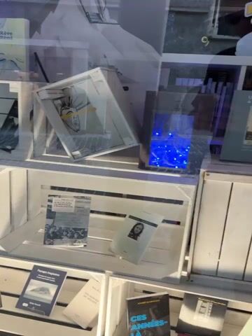 The video shows a display of books in a store window. The books are arranged on white wooden crates. In the top shelf, there is a book with a blue cover titled "Le Rêve de Rémi." To the left of this book is a book with a red cover titled "FRISSONS D'ÉTE." In the middle shelf, there is a book with a black cover and another book with a white cover. In the bottom shelf, there are several books with various titles and colors. Some of the books are standing upright, while others are lying flat. The display is well-lit, and the books are clearly visible. The overall impression is one of a carefully curated book display.