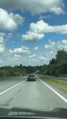 The video shows a view from the front of a vehicle driving on a highway. In the distance, another car is traveling in the same direction. The highway is asphalt with white lane markings. On either side of the road, there are green trees and vegetation. The sky is blue with scattered white clouds. The scene appears to be a daytime shot of a rural landscape during a drive.