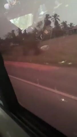 The video shows a view from inside a moving vehicle, looking out the window at a rural landscape. The road is reddish-brown. Trees and buildings are visible in the background. The window has some smudges and water spots on it. The video is shaky due to the movement of the vehicle. Someone speaks in the background.