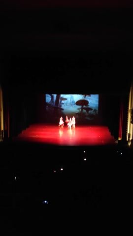 A group of six dancers in white costumes perform on a dimly lit stage. The dancers are arranged in two rows, with three in the front and three in the back. They are all facing the audience and appear to be in the midst of a choreographed routine. Behind them, a large screen displays a colorful animation of what appears to be a whimsical landscape with oversized mushrooms. Red stage lights illuminate the dancers and create a hazy atmosphere. The overall scene suggests a performance, possibly a ballet or a theatrical production, for a young audience.
