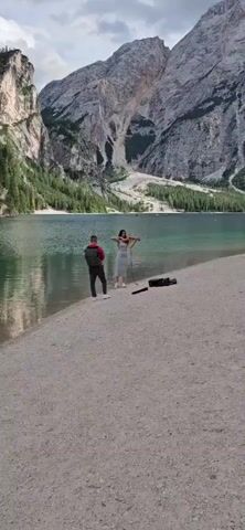 A man stands on a sandy shore, his back to the camera, facing a woman who is playing a violin. The woman is wearing a light-colored dress. A violin case and a microphone are on the ground near her. Behind them is a large body of water, likely a lake, with mountains and trees in the background. Several boats are visible on the water. The scene appears to be outdoors in a natural setting.