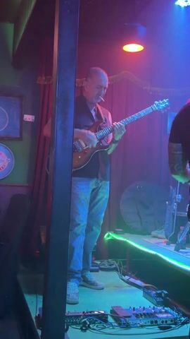 The video shows a man playing an electric guitar in what appears to be a dimly lit bar or music venue. The man is standing on stage, wearing a black vest, light blue jeans, and grey shoes. He has a cigarette in his mouth. He is playing the guitar with focus and expression. The stage is decorated with green neon lights along the front. Behind him is a dark red curtain. To his left is a large metal post, slightly obscuring the view. There are various pieces of equipment on the floor, including a pedalboard. Another person, partially visible, is standing to the right of the guitarist. The room has a dim, reddish-purple hue with additional lighting accents. A framed image is seen on the wall to the left. The atmosphere suggests a live music performance in a casual setting.