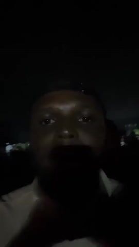 The video is taken at night and is mostly dark. At the beginning, a blurry figure that could be a person is in the foreground. The person is slowly revealed by the camera's light, showing a person with dark skin wearing a dark baseball cap. They appear to be at an outdoor event, with other people visible in the background. The illumination is very poor, but it appears that the people in the background are also attending the same event. There is music playing in the background.