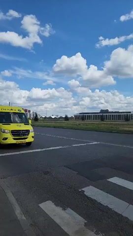 A yellow Mercedes-Benz Sprinter van with the text "По пути" on its side drives across a pedestrian crossing. The van is yellow with some blue and white graphics. In the background, there are modern buildings, a blue sky with white clouds, and a road with another yellow vehicle driving away. A person stands on the pedestrian crossing, looking at their phone. The scene appears to be on a sunny day in a modern urban or suburban environment.