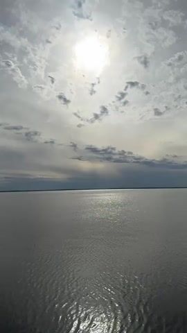 The video starts with a wide shot of a vast, calm body of water under a cloudy sky where the sun shines through, creating a bright glare on the water's surface. The camera angle shifts down to show a concrete edge or embankment where the water meets the land. The camera then focuses on the gray, cracked concrete surface, before revealing two pairs of legs and shoes standing on it. One pair of feet is wearing white sneakers and gray pants, while the other pair is wearing light brown sneakers and light blue pants.  No action or interaction is occurring between the individuals. The environment is serene but with visible signs of wear and tear on the concrete structure.