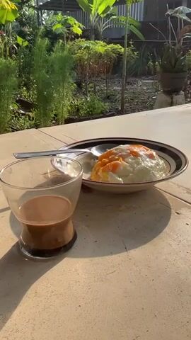 A table with a meal of rice and eggs and a glass of coffee. The table is outdoors, with plants and a building in the background. The sun is shining.