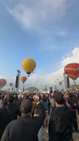 The video shows a large crowd of people gathered at what appears to be an outdoor event. People are standing and looking up, many holding up their cell phones to record or take pictures. Numerous hot air balloons are in the air, including yellow, red, and multicolored ones. The sky is blue with scattered clouds. In the background, there is a pyramid-like structure, likely a significant landmark. The crowd is surrounded by tall speaker towers. The audio includes music with a strong beat.