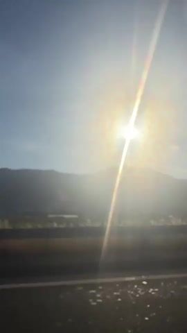The video shows a bright, sunlit sky with a mountain range in the background. The sun is shining directly into the camera lens, creating a strong glare and light streaks. A road or other flat surface is visible in the foreground. The sound is dominated by strong wind noise.