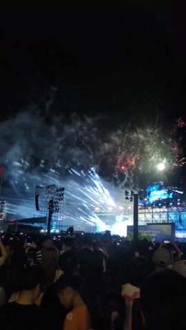 The video captures a nighttime concert or festival scene. A large crowd is gathered facing a stage illuminated by bright white spotlights. Fireworks explode in the sky, visible above the stage and crowd. Two large screens flank the center stage, displaying what appears to be a performer. Many in the audience are holding up their phones, presumably recording the event. Banners and signage with logos are visible on the stage. The sky is dark, with smoke from the fireworks drifting. The scene suggests a celebratory atmosphere at a large outdoor event.