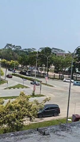 A horizontal video shows a road intersection with trees and buildings in the background. Several cars are visible on the road, including a black SUV parked on the right side of the screen. There are also lampposts and traffic signs. The sky is clear and blue. The video is shaky, suggesting it might have been recorded by hand.