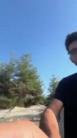 Here is a description of the video:

The video begins with a selfie shot of a young man with short, curly dark hair, a beard, and glasses, wearing a black t-shirt. He's standing outdoors on a gravelly patch of ground.  Behind him, there are several large tank trucks.

The camera pans up to show a wider view. A man is on top of one of the trucks. Other trucks are visible in the background, along with trees and power lines against a bright blue sky with scattered clouds. Someone walks towards the left side of the frame.