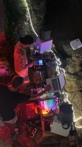 A top-down, wide shot shows two people setting up a DJ station outdoors at night. The DJ station is comprised of multiple electronic music devices including mixers, a laptop, speakers, and a keyboard, all connected by wires and illuminated by colorful lights. The two individuals appear to be working together, adjusting the equipment. The environment is a rough, outdoor setting with stone walls and vegetation. In the background, a building and a distant cityscape with lights are visible, suggesting an urban or semi-urban location at dusk or night. A white string of lights zigzags across the scene, adding to the ambient lighting.