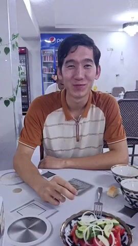 A man with dark hair, wearing a brown and cream striped shirt, sits at a table in a restaurant. He smiles and waves at the camera at 00:01. In front of him on the table are plates, bowls, cutlery, a bread roll, and a salad. Behind him, a Pepsi refrigerator is visible, and other patrons are in the background. At 00:04, the camera pans to show more of the restaurant, including tables with checkered chairs and a menu displaying food items. The man continues to smile.