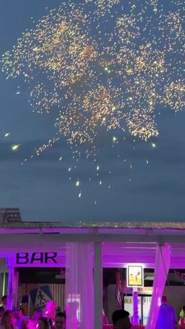 This media file captures a fireworks display over a beach bar at dusk in Дрокино, Russia. The sky is a deep twilight blue, with scattered clouds. Numerous fireworks of varying sizes and colors, predominantly gold and yellow, burst across the sky, illuminating the scene.

Below the fireworks, a white-roofed beach bar is visible, lit with vibrant purple lights. The bar has "BAR" written in large white letters on its facade. Sheer white curtains hang from the structure, adding a soft, ethereal glow. People are gathered at the bar and around the area, their silhouettes visible against the illuminated interior. One individual in the foreground is out of focus.

The event appears to be a celebration or festive occasion, with the fireworks serving as a spectacle for the patrons of the beach bar. The atmosphere seems lively and festive, given the celebratory nature of fireworks. The time of day, dusk, suggests the event is taking place in the early evening.
