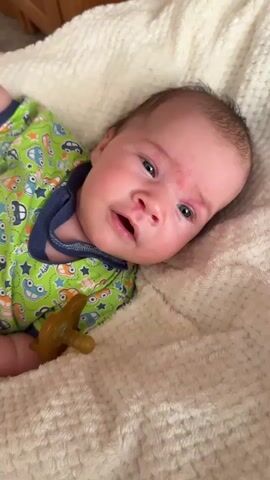 A close-up video shows a newborn baby lying on its back on a cream-colored, textured blanket. The baby is wearing a green onesie with a repeating pattern of colorful cars. A tan pacifier rests near the baby's right side. The baby's head is tilted slightly to the right. The baby's eyes are open and looking upwards. The baby's mouth is open slightly, and they appear to be making a soft vocalization. The baby's skin is slightly flushed, particularly on the cheeks and around the eyes. The lighting suggests an indoor environment. The baby shifts slightly, and their eyes close briefly before opening again. The baby then turns their head slightly to the left, and their facial expression changes, appearing to be distressed or crying. Their eyes are closed, and their mouth is open. The baby's body remains mostly still.