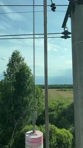 The video captures a view from a moving vehicle, likely a train, looking out at a landscape. Overhead power lines run horizontally across the frame. The scene is dominated by lush green trees and foliage. In the background, a large, open field or meadow is visible, bordered by a dense line of trees. The sky is a pale blue with scattered white clouds. The lighting suggests it is daytime, possibly late morning or early afternoon. There are no people or animals visible in the video. The perspective suggests the viewer is stationary, looking outward.