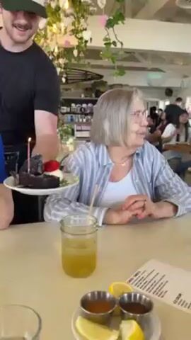 A woman sitting at a counter receives a small birthday cake from a waiter. The cake is a square of dark chocolate cake with a single lit candle, dusted with powdered sugar and topped with a strawberry and a dollop of whipped cream. The woman is wearing a white shirt and a blue and white striped button-down. She has short gray hair and glasses. There is a jar of yellow liquid and a slice of lemon on the counter in front of her. A man in a black shirt and apron is holding the cake in front of her. Another person is wearing a blue shirt. Other patrons are visible in the background of the bright, airy cafe. The woman smiles widely as the cake is presented. Someone sings "Happy Birthday".