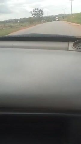 The video shows a view from inside a vehicle, looking out through the windshield. The dashboard is visible in the foreground. A road stretches ahead, with some trees and vegetation on either side. There are a few vehicles on the road, including a bus in the distance. The sky is overcast. The video appears to be a first-person perspective of someone driving or riding in a car.