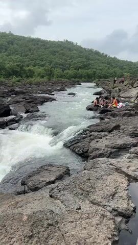 The video shows a view of a river with rapids flowing over dark rocks. In the background, there is a lush green hill covered in trees, with a cloudy sky overhead. On the right side of the riverbank, a group of people are sitting on the rocks. The camera pans slightly to the right, showing more of the rocky riverbed and the flowing water. The video appears to be taken during the day, likely in a natural, outdoor setting.