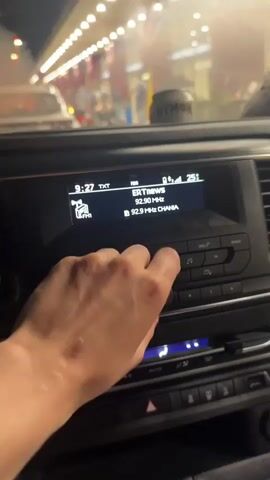 A person's hand is shown in a car, adjusting the volume on the car's display screen. The screen reads "Volume" and shows a bar indicating the volume level, which is set to "10." The radio station is "ERT MWE" at "92.50 MHz," also showing "92.9 MHz CHANIA." In the background, there's a blurry view of a tunnel or an indoor parking lot with bright overhead lights. A light-colored can with the word "NYMOH" is visible on the dashboard.
