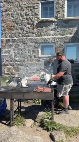 The video shows an outdoor cooking scene with a group of people. In the foreground, a man with a beard wearing a black t-shirt and shorts is tending to a grill with sausages. He is wearing black gloves. To his left, another man in a white t-shirt is serving food from a metal container. A blue crate is visible on the ground next to him. Behind the grill, there are several people in line, some wearing backpacks, suggesting they are hikers. The background features a stone building with windows and a clear blue sky. The environment appears to be a mountainous or elevated area, with vegetation and rocky terrain visible. The overall atmosphere suggests a casual outdoor meal or gathering.