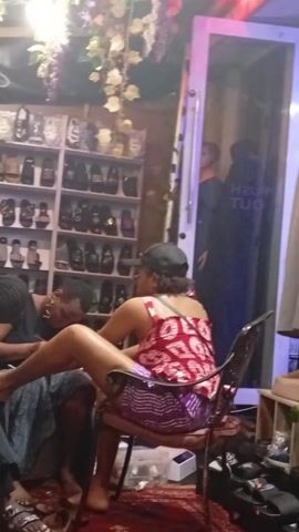 The video shows the interior of what appears to be a salon or retail store. There are two women present. One woman, seated in a chair, is attending to the feet of the other woman, possibly giving her a pedicure. A large shelf displays numerous pairs of sandals. A glass door with "PUSH OUT" visible is behind the seated woman. The environment is somewhat cluttered with various objects and décor.