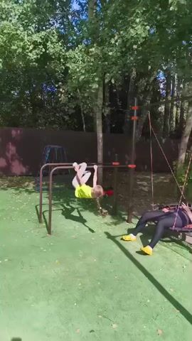 A young child is performing a gymnastic move on a horizontal bar, hanging upside down with their legs bent. They are wearing a bright yellow t-shirt and grey leggings. To the right, a woman is lying on a circular swing seat, her head tilted back and eyes closed, appearing relaxed. She is wearing black leggings and a grey top, with bright yellow shoes. A small dog is lying on the green, artificial turf in the foreground, facing left. The background consists of trees with dense green foliage, a brown fence, and other playground equipment, including a blue and red climbing structure and more horizontal bars. The lighting suggests it is a sunny day.