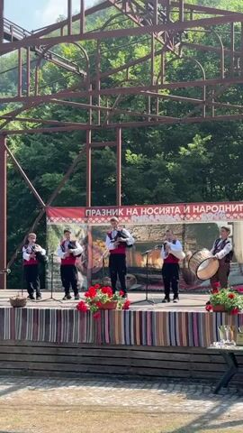 A group of five young males are performing on an outdoor stage. They are dressed in traditional Bulgarian folk costumes, consisting of white shirts, red vests, and dark pants. All five are playing musical instruments, specifically bagpipes or a similar wind instrument. The stage is decorated with a colorful striped rug and red flowers in pots. Behind the performers, a banner with Bulgarian text and imagery is visible. The stage is supported by a metal structure, and a dense forest can be seen in the background. The lighting suggests it is daytime, with sunlight illuminating the scene.