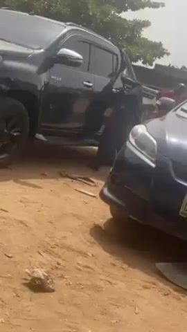 The video shows a black Toyota Hilux parked on a dirt ground next to a blue sedan, with a person standing in between the two vehicles. The person is wearing dark clothing and sandals and is standing near the front of the blue car. The ground is sandy and there are some small pieces of debris scattered around. There are trees visible in the background.