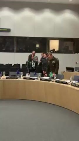 Here is a detailed description of the video:

The video shows an indoor meeting taking place in what appears to be a conference room. The room is spacious and features a large, curved table made of light-colored wood, around which chairs are arranged. At the table, several individuals are standing and interacting.

The individuals are dressed in a variety of professional attire including military uniforms. There are at least two people in military uniforms, one of whom is standing towards the right side of the frame, apparently looking at documents on the table. A woman wearing a green and black plaid jacket is standing towards the left of the frame. A man in a dark suit is walking away from the group toward the back of the room.

The table is set with water bottles, small flags (likely representing countries or organizations), and papers. There are individual nameplates at each seating position on the table.

Behind the table, there is a large window with multiple panes, and reflections of the room and its occupants can be seen in the glass. In the very back of the room, empty chairs can be seen.

The camera is static, and the lighting is bright, indicating a well-lit indoor environment. The overall scene appears formal and professional, suggestive of an official meeting or gathering.