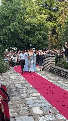 A woman in a long, light blue dress walks down a red carpet with a man, followed by a child. The carpet is lined with rose petals. A crowd of people watches from behind a barrier to the left of the carpet. The setting appears to be outdoors, with trees and greenery in the background.