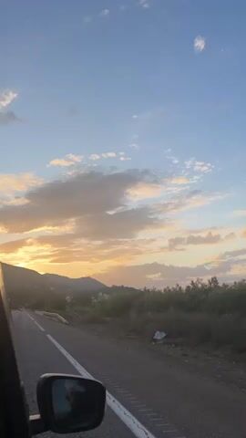 From the perspective of the inside of a vehicle, the video shows a drive on a road or highway at dusk.  The car's side mirror is visible in the lower left of the frame. The road has a white line on the left and a gray cement barrier with blue accents on the right. In the distance are mountains partially silhouetted by the setting sun, with scattered buildings and trees dotting the landscape.  The sky is a blend of light blue and golden hues with a variety of cloud formations. There is light traffic on the road ahead.