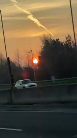 The video shows a sunset from the perspective of someone driving on a highway. The sun is low on the horizon, appearing orange and partially obscured by trees. A long contrail stretches across the sky. The highway has a concrete barrier along the side, and there are streetlights lining the road. The overall tone of the video is peaceful and serene.