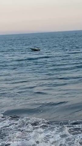 A video captures a wide view of a body of water, likely the ocean, under a hazy sky. In the distance, a small boat with a few people on board moves across the waves. The foreground shows the crest of a wave breaking onto the shore, creating white foam. The overall scene is serene, with the gentle movement of the water and the distant boat creating a sense of peace. The video is brief, only a few seconds long, and the camera is stationary, capturing the natural beauty of the coastal environment.