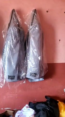 The video shows the interior of what appears to be a tailoring or sewing workshop. Three gray suits covered in clear plastic hang on a red and white wall. Below them, on the floor, are folded pieces of clothing and a cardboard box. The camera pans right, revealing a sewing machine on a white table. A mirror leans against the wall nearby. The workshop seems small and cluttered.