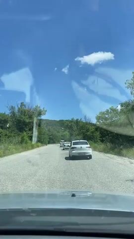 The video shows a view from inside a vehicle driving on a road, likely in Татарка, Russia. The road is asphalt and appears to be in fair condition, with some cracks visible. It is a sunny day with a clear blue sky and fluffy white clouds. Lush green trees and vegetation line both sides of the road, and in the distance, rolling hills are visible, covered in more greenery.

Several white cars are driving ahead of the vehicle, forming a small convoy. The closest car to the camera is a white sedan, which is the primary subject in the immediate foreground of the road. Further ahead, a white van or minibus and another car are also visible.

The scene suggests a peaceful drive through a rural or semi-rural area during the daytime. The bright sunlight and clear skies indicate pleasant weather. There is no visible text within the video frame itself. The overall mood is serene and relaxed, as one might experience on a road trip or scenic drive. The perspective from the driver's seat adds a sense of immediacy and immersion.