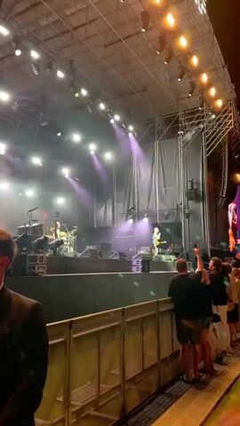 A group of people are watching a band perform on an outdoor stage at night. The stage is lit with bright lights, and smoke or fog is being used for visual effects. A guitarist is in the center of the stage, playing a guitar and singing. The audience is standing behind a metal railing, some are holding up their phones to record the performance.