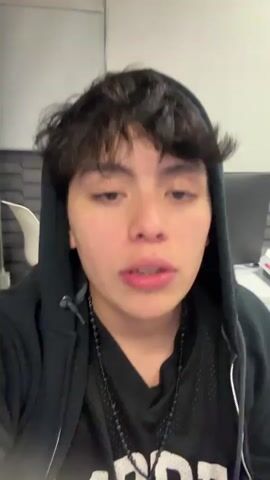 A young person wearing a black hoodie and a beaded necklace is looking at the camera and talking. They rub their eye with their fingers. In the background is an office environment with a woman sitting at a desk working on a Dell computer. Upbeat music plays.