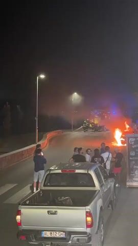 A nighttime scene features a vehicle fire on a road. Several firefighters in yellow helmets and dark uniforms are visible near the burning vehicle, which is engulfed in flames and smoke. Red and blue lights from emergency vehicles illuminate the area. A group of onlookers, some holding cell phones, are standing on the side of the road, observing the incident. In the foreground, the back of a silver pickup truck is visible, parked on a crosswalk. The environment appears to be an outdoor setting, possibly near a roadside establishment, with streetlights casting illumination.