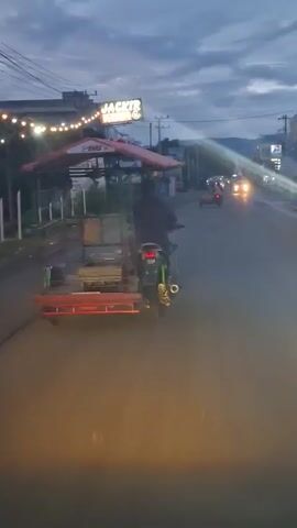 A person drives a motorcycle with a red-canopied sidecar attached to its left, moving away from the camera on a paved road during twilight. The sidecar is red with green metal framing and a red canopy displaying an "MB" logo. Various items, including what appears to be cooking equipment, are visible in the sidecar. The driver is wearing a dark shirt. On the left side of the road, there are buildings, one with a "JACKTR Kopi" sign, and illuminated string lights. Power lines and utility poles run along the left side. Distant mountains are visible on the right horizon. Other vehicles, with their headlights on, are present further down the road. The sky is a cloudy blue-grey.