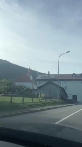 The video shows a view from inside a vehicle looking out at a rural village scene. The foreground shows the dashboard and windshield of the vehicle, with windshield wipers visible. In the middle ground, a paved road runs from right to left. To the left of the road is a grassy hill with a small building with a red roof and a steeple. Further to the right is a larger building with a light-colored facade and a dark roof. A street lamp stands in front of the larger building. On the right side of the road, a motorcycle with two riders passes by. The sky above is clear with a hazy sun.