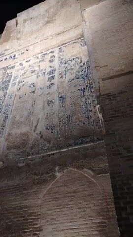 A low-angle, handheld shot pans upwards, revealing a section of a weathered, ancient wall. The wall is constructed of rough, aged stone or stucco, with visible patches of deterioration and discoloration. Dominating the upper portion of the wall are numerous rectangular panels adorned with intricate, faded blue tilework. These tiles appear to be arranged in vertical columns with decorative elements and possibly Arabic script. The lower part of the wall transitions to exposed, rough-hewn brickwork, forming an arched opening or alcove. The lighting is dim, suggesting it is nighttime or an interior space with limited illumination, casting shadows and highlighting the texture of the materials. The overall impression is one of historical significance and age.