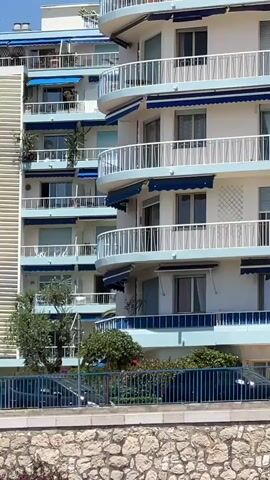 The video shows a multi-story apartment building with balconies. The building is white with light blue trim and blue awnings on the balconies. There are some trees and plants in front of the building, and a stone wall in the foreground. A few people are visible on the balconies and walking on a sidewalk in front of the building. The sky is clear and blue.