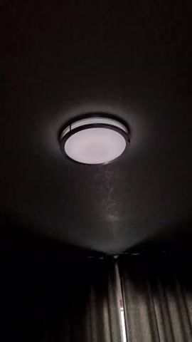 This is a close-up, low-angle video of a ceiling light fixture in a dimly lit room. The fixture is circular with a metallic rim and a white diffused lens. The light flickers on and off, cycling through dimmer and brighter states. Gray curtains are visible at the bottom of the frame. There are no people or other discernible objects in the video.