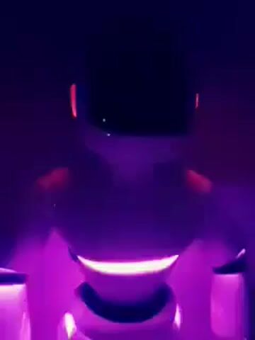 A stationary, humanoid-shaped robot is depicted in a dark setting, illuminated primarily by its internal LED lights. The robot's body is light-colored, featuring a prominent band of lights around its midsection. These lights dynamically cycle through various colors, including bright blue, vibrant red, and a warm yellow/orange hue, creating a striking visual contrast against the dark background. The robot's head appears dark, with only a subtle light visible near the top. The video focuses closely on the robot's upper body, showcasing the mesmerizing color changes of its integrated lighting system. No other objects or text are visible, emphasizing the futuristic and technological aesthetic of the robot.